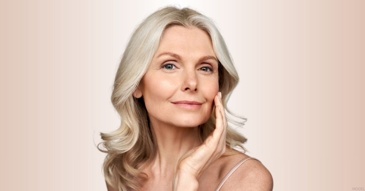 Woman with shoulder-length gray hair touches her face on a beige background. (model)
