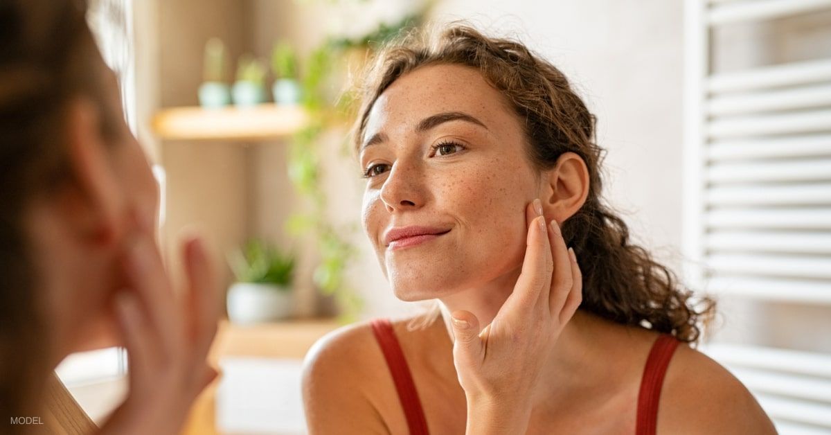 Woman with clear and radiant skin (model) looking in the mirror while touching her face.